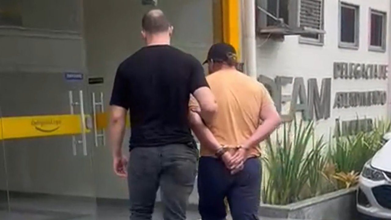 Homem algemado sendo escoltado por policial em frente à delegacia DEAM-NF.