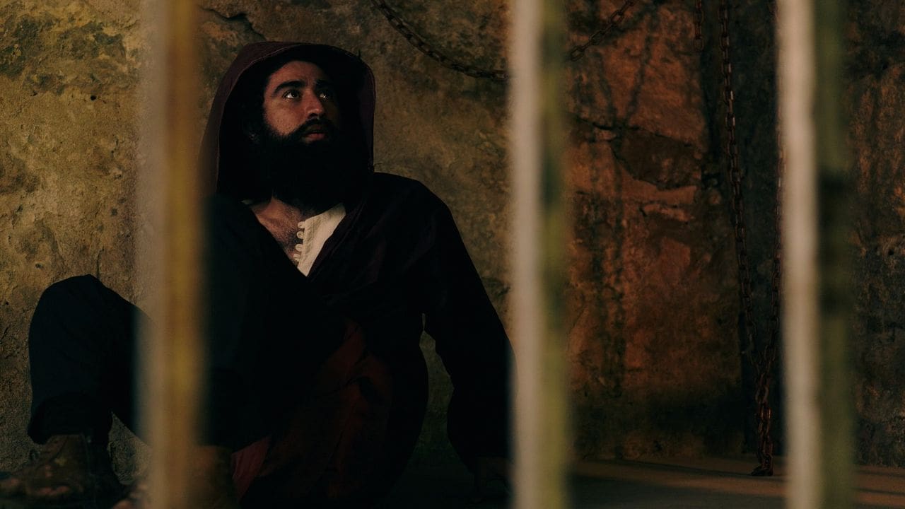 Homem com barba e capuz em ambiente histórico, possivelmente uma prisão ou masmorra.