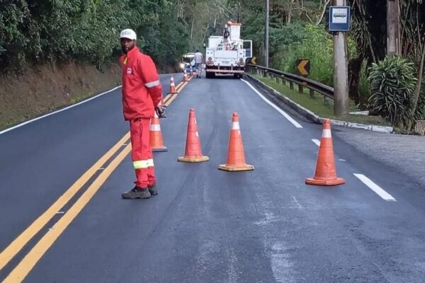 Homem em uniforme laranja em serviço de manutenção rodoviária, com cones de sinalização e veículo de trabalho.