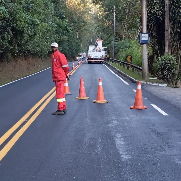 Homem em uniforme laranja em serviço de manutenção rodoviária, com cones de sinalização e veículo de trabalho.