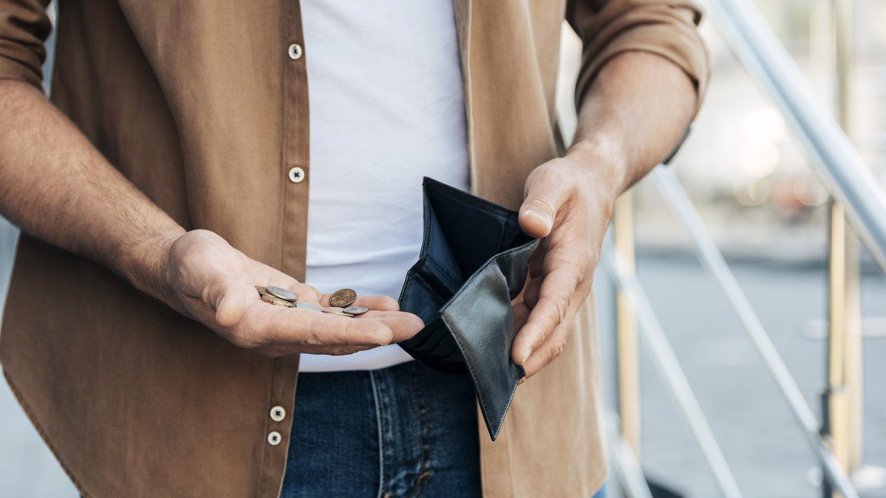 Homem mostrando carteira vazia e algumas moedas na mão, simbolizando dificuldades financeiras.