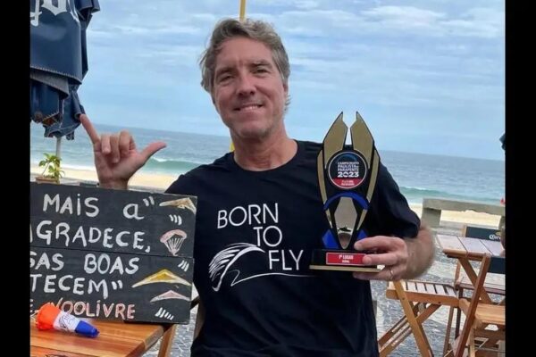 Homem segura troféu de voo livre com a frase 'Born to Fly' na camiseta.