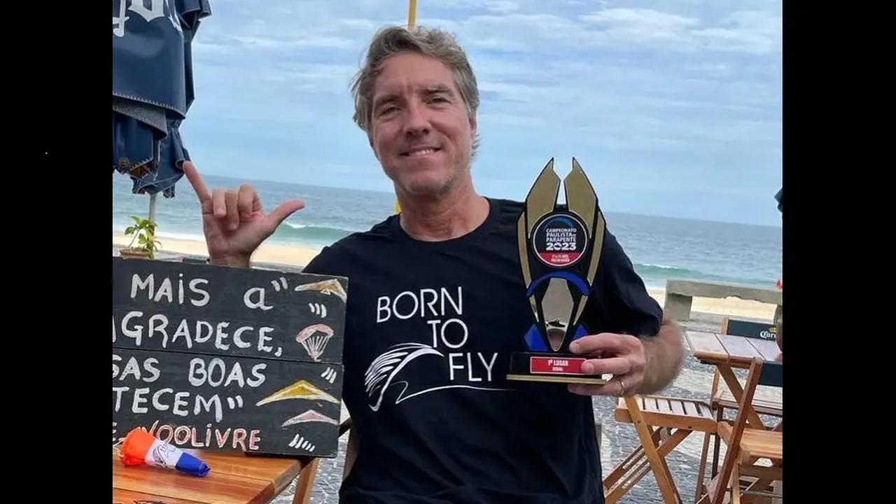 Homem segura troféu de voo livre com a frase 'Born to Fly' na camiseta.