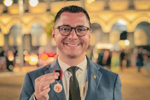 Homem sorrindo com medalha em frente a prédio histórico.