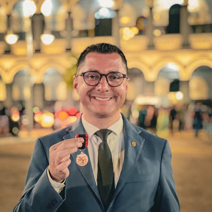 Homem sorrindo com medalha em frente a prédio histórico.