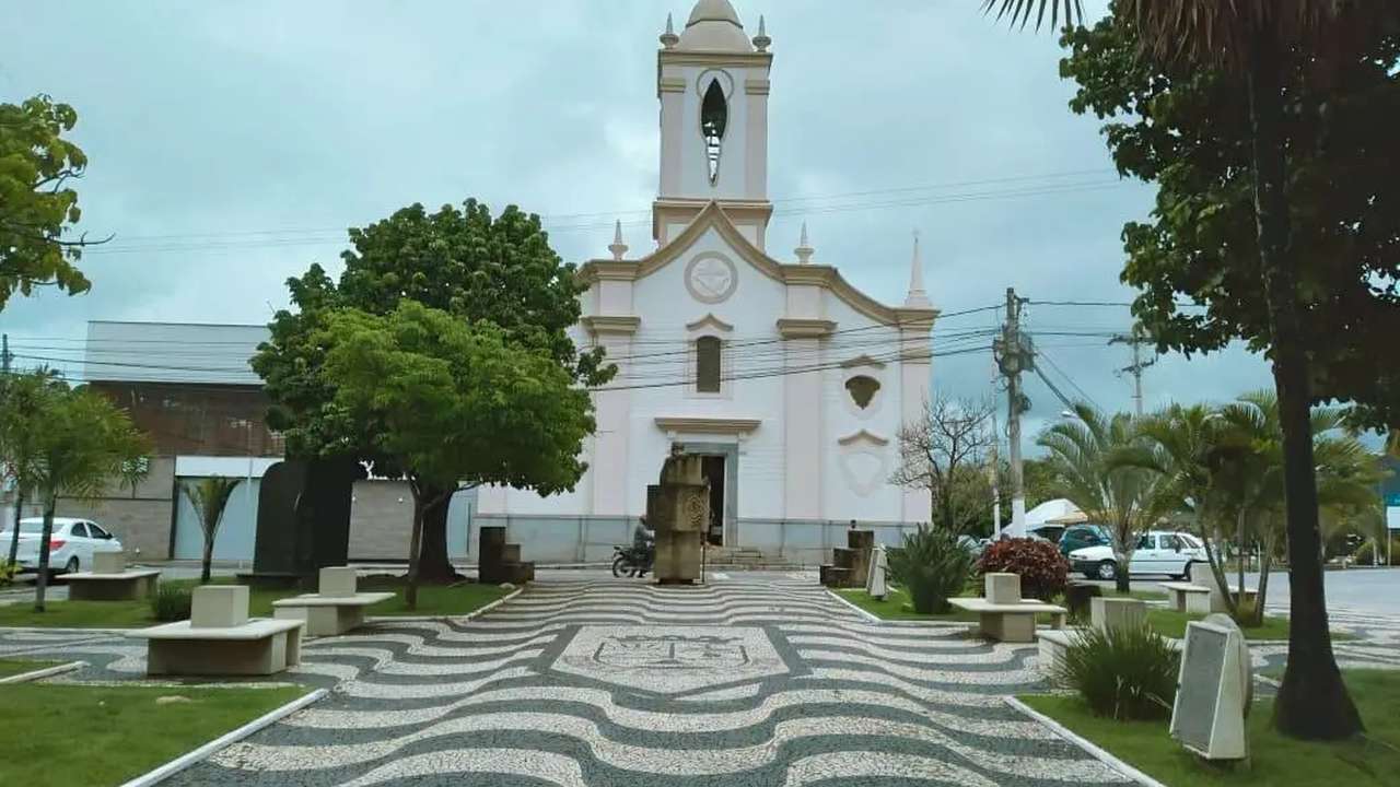 Igreja em Itaocara, Rio de Janeiro, com calçada em padrão ondulado e árvores ao redor.
