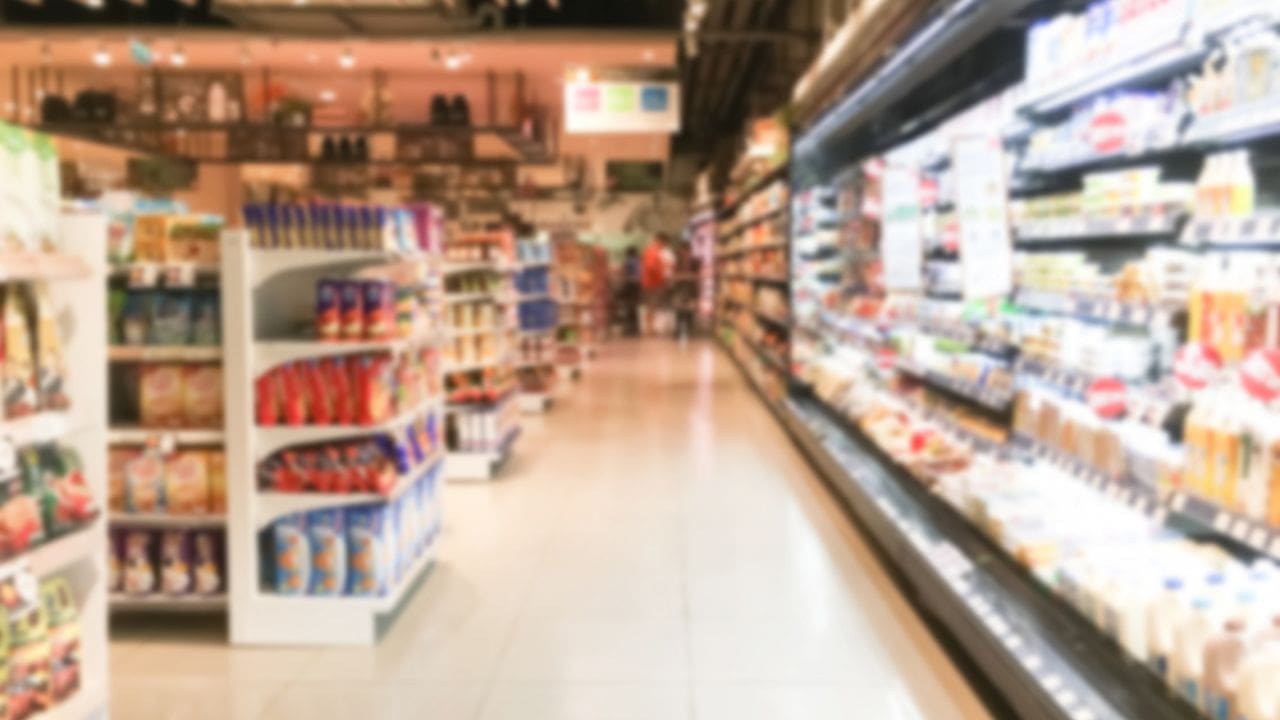 Imagem borrada do interior de um supermercado, corredores com prateleiras de produtos e pessoas ao fundo.