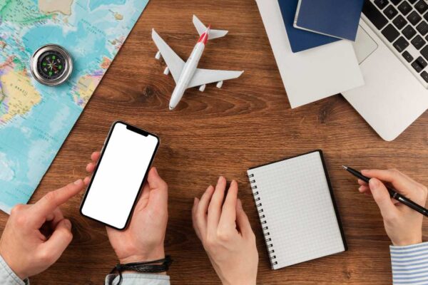 Imagem de uma mesa com mapa, smartphone, avião de brinquedo, caderno, caneta e laptop, simbolizando planejamento de viagem.