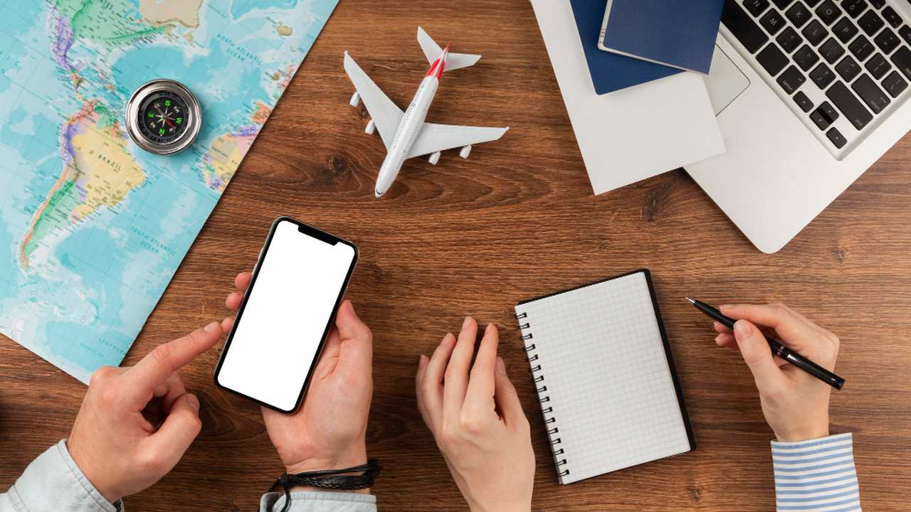Imagem de uma mesa com mapa, smartphone, avião de brinquedo, caderno, caneta e laptop, simbolizando planejamento de viagem.