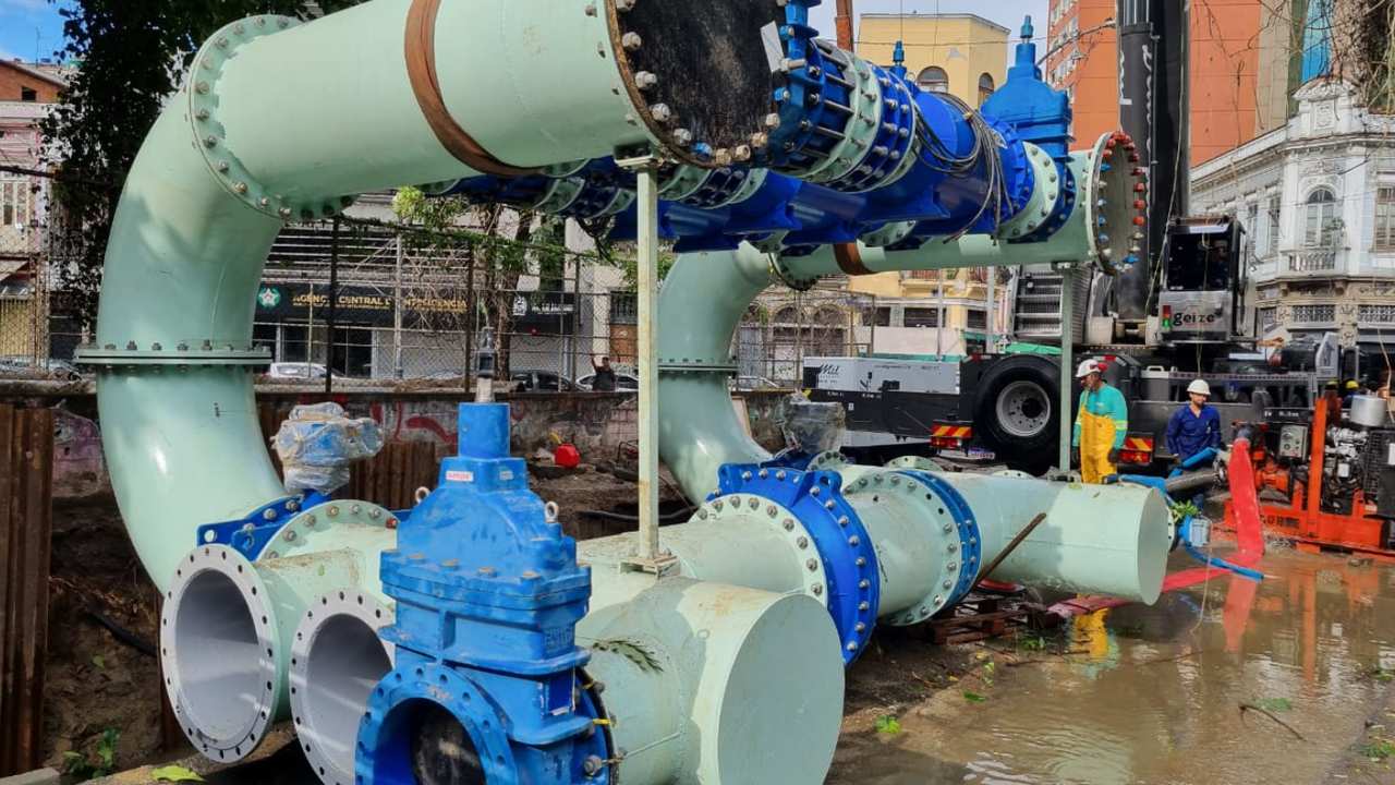 Instalação de tubulação em obra de saneamento, com trabalhadores e guindaste.