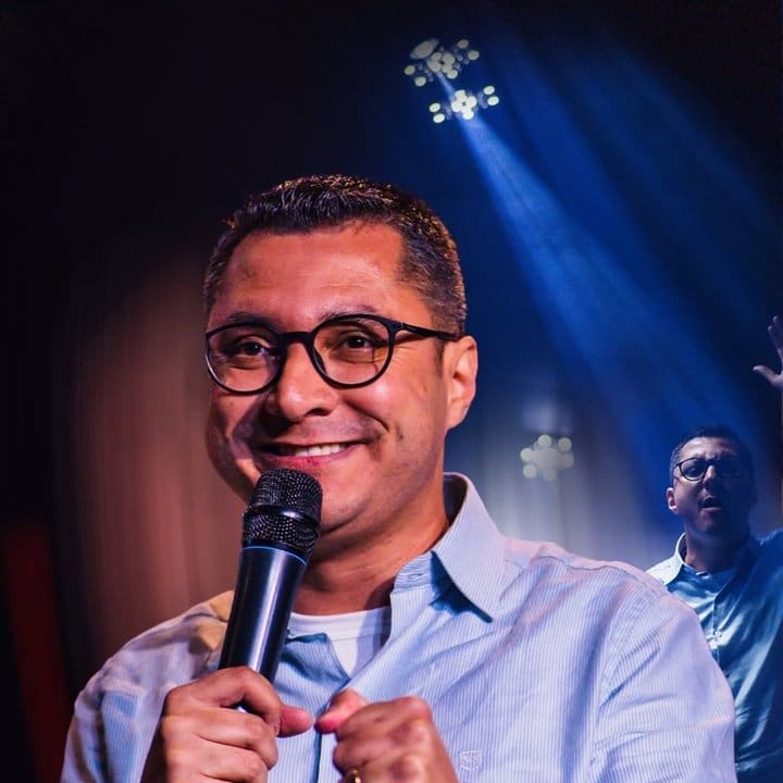 Johnny Maycon, prefeito de Nova Friburgo, em evento público, com microfone e óculos.