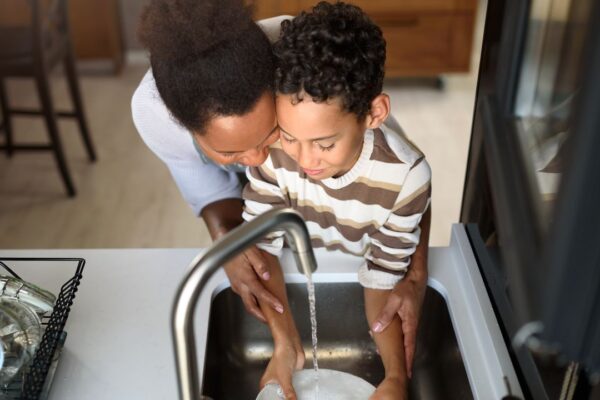 Mãe e filho lavando louça na pia da cozinha, com foco em conscientização sobre água.
