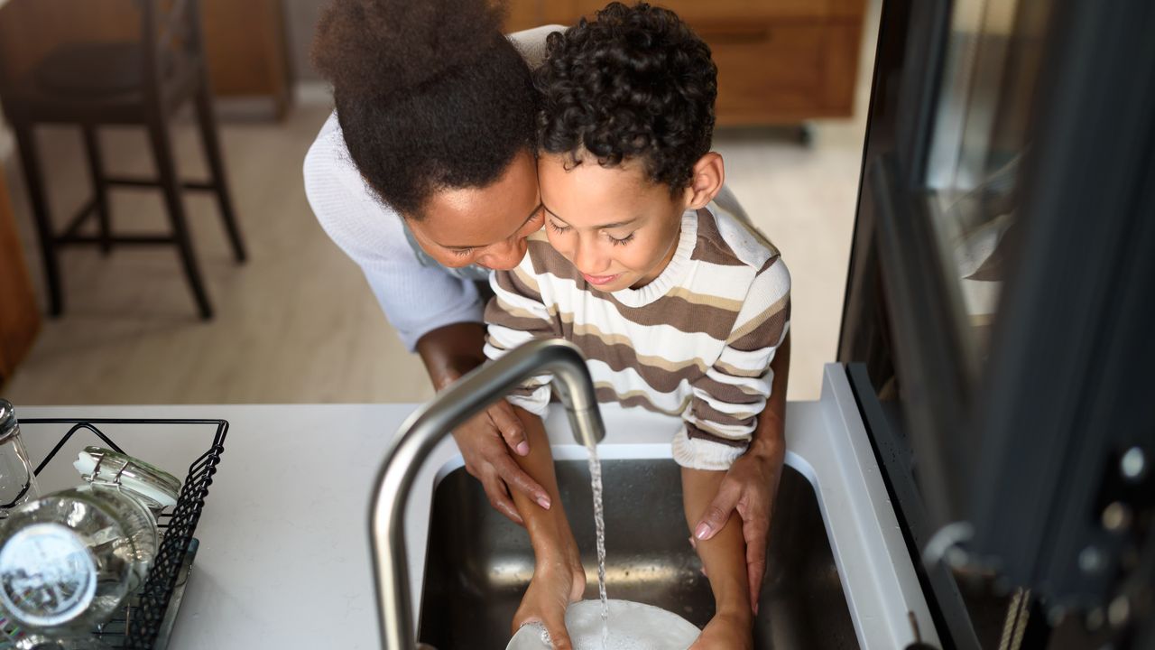 Mãe e filho lavando louça na pia da cozinha, com foco em conscientização sobre água.
