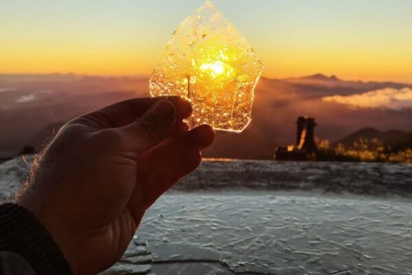 Mão segura pedaço de gelo com o sol brilhando por trás, no Pico do Caledônia congelado.