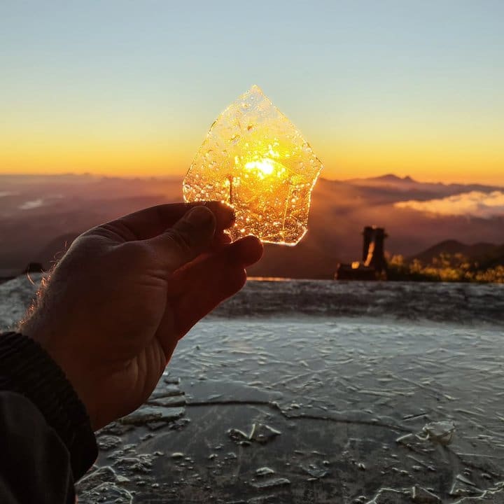 Mão segura pedaço de gelo com o sol brilhando por trás, no Pico do Caledônia congelado.