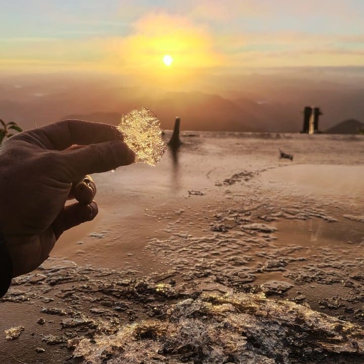 Mão segurando gelo refletindo a luz do sol, com paisagem montanhosa ao fundo.