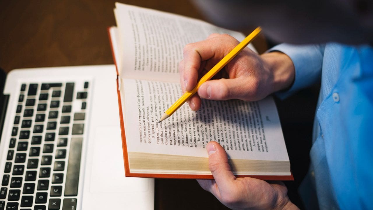 Mãos segurando livro aberto e lápis, com teclado de notebook ao fundo