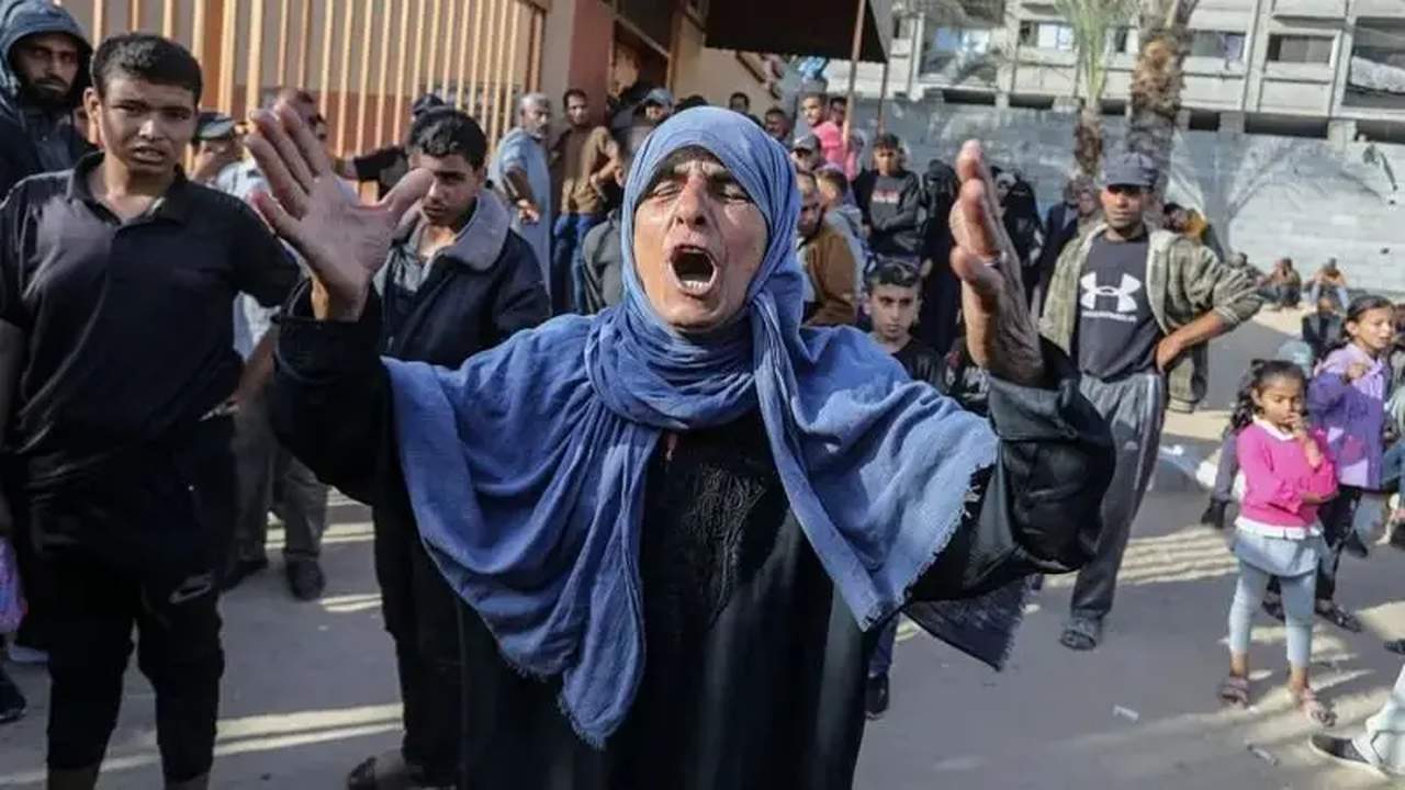 Mulher em desespero durante ataque israelense, com braços erguidos e multidão ao fundo.