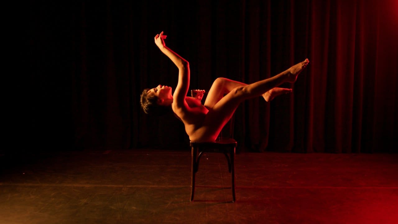 Mulher nua em pose artística em palco com iluminação dramática.