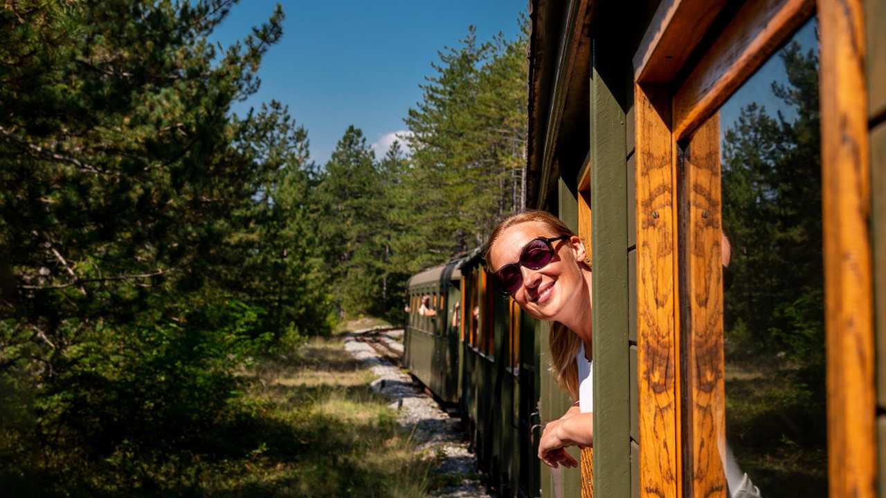 Mulher sorrindo na janela de um trem, com óculos escuros, em meio à paisagem.