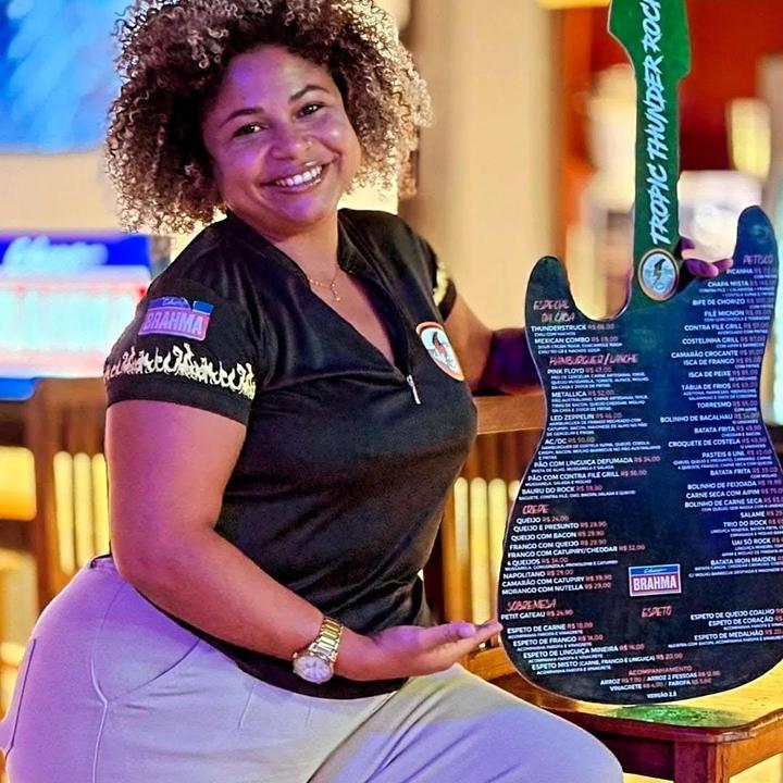 Mulher sorrindo segura cardápio de restaurante 'Tropic Thunder Rock' em formato de guitarra.