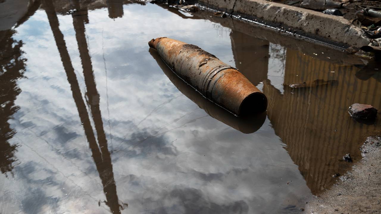 Objeto enferrujado em água refletindo o céu e estruturas.