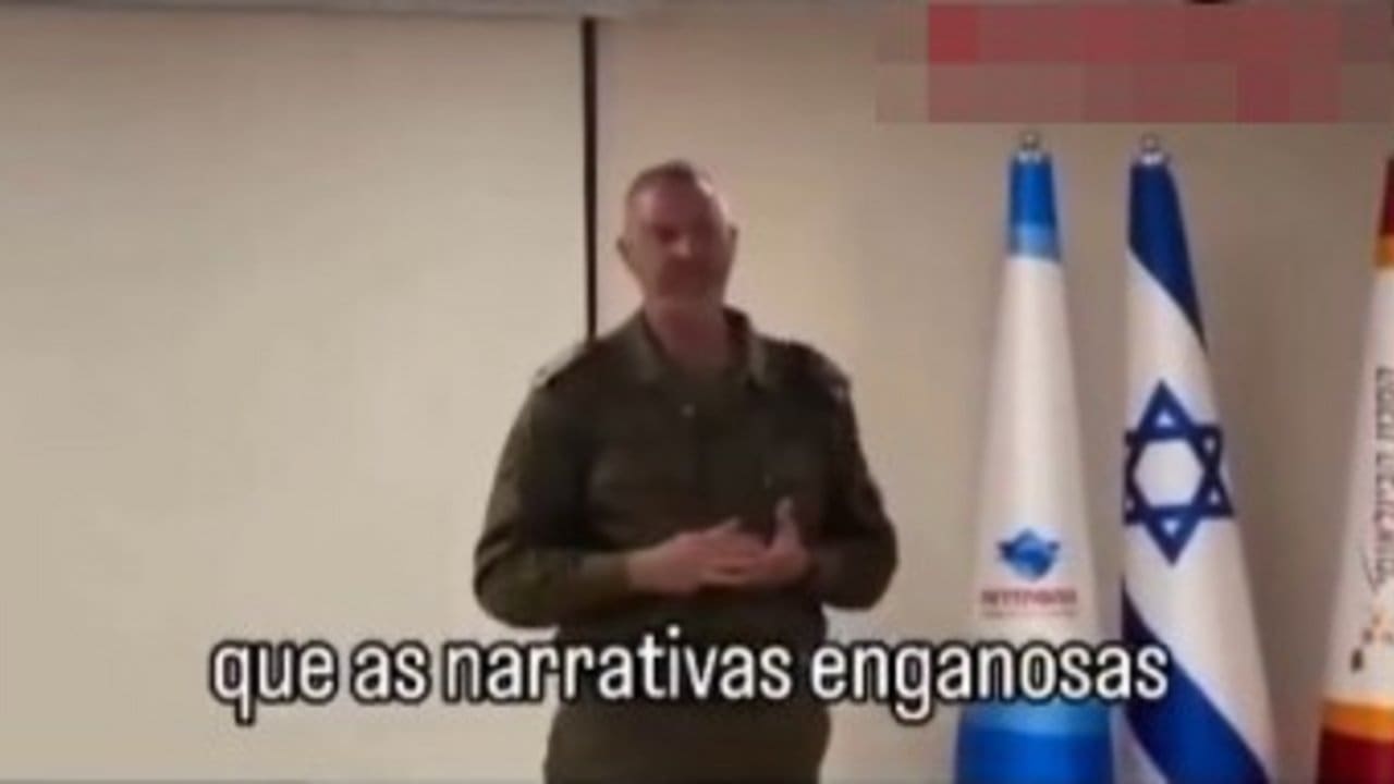 Oficial israelense em apresentação sobre narrativas enganosas, com bandeiras de Israel ao fundo.