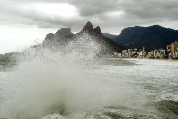Ondas fortes quebrando na praia com montanhas ao fundo e cidade costeira sob céu nublado.