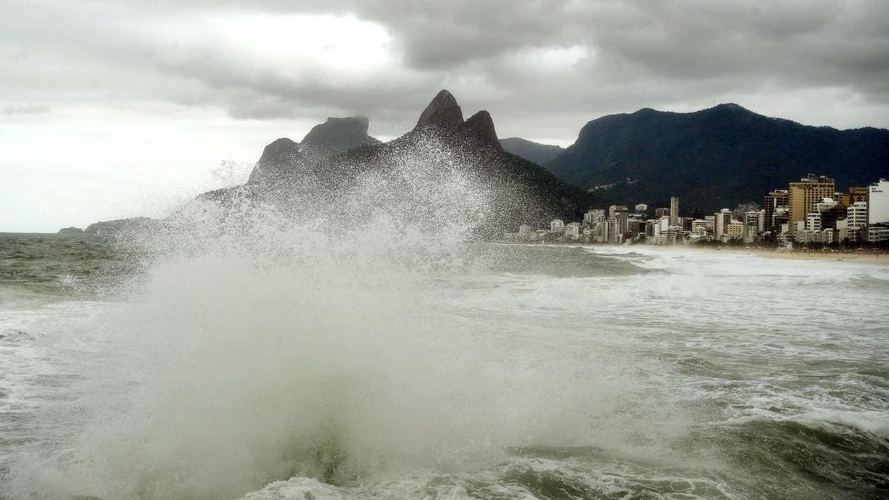Ondas fortes quebrando na praia com montanhas ao fundo e cidade costeira sob céu nublado.