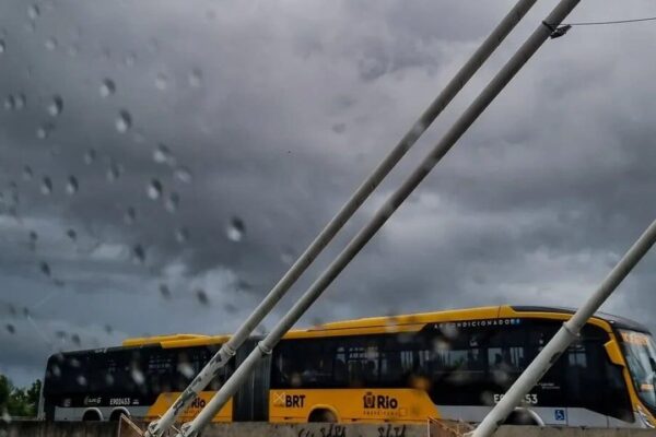 Ônibus amarelo em uma parada de ônibus, com chuva caindo e céu nublado.
