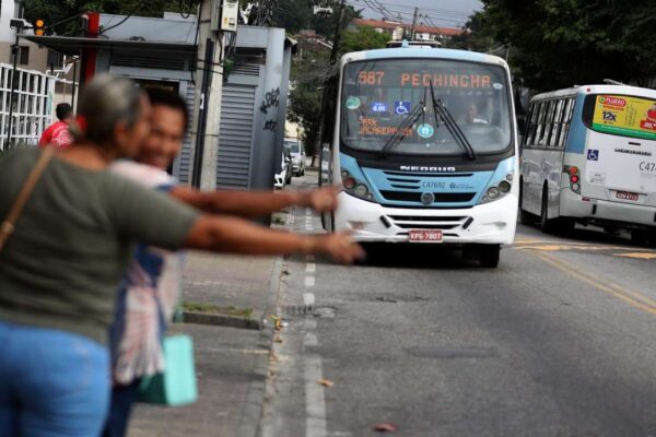 Ônibus de transporte público em via urbana, com pessoas no primeiro plano tentando sinalizar para o veículo.