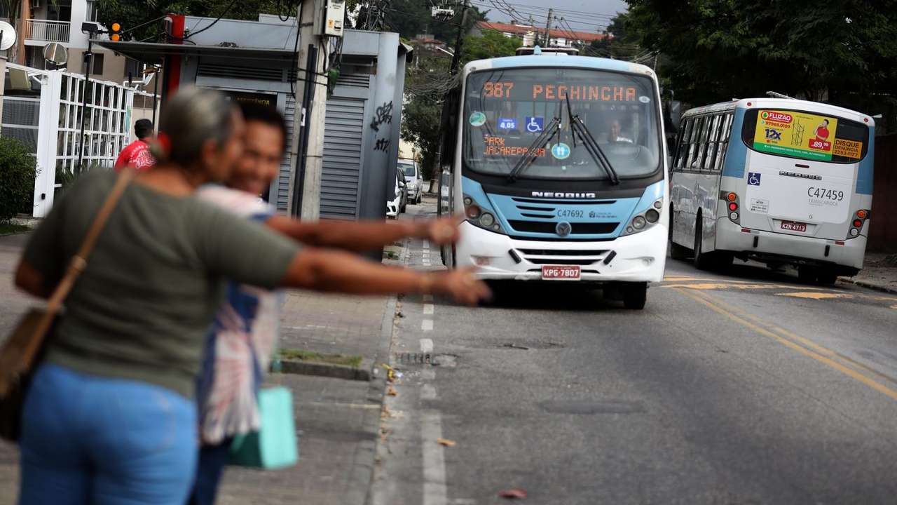 Ônibus de transporte público em via urbana, com pessoas no primeiro plano tentando sinalizar para o veículo.