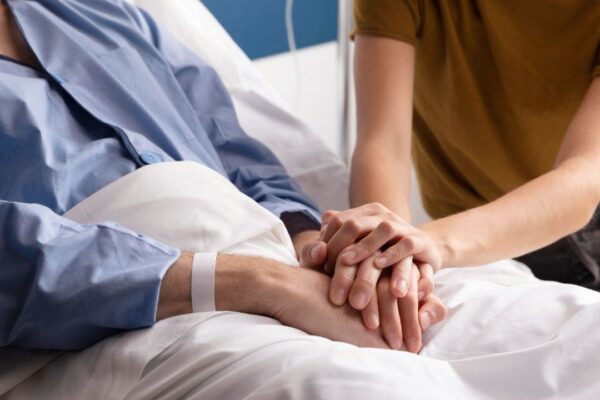 Paciente em leito hospitalar com alguém segurando suas mãos em sinal de apoio.