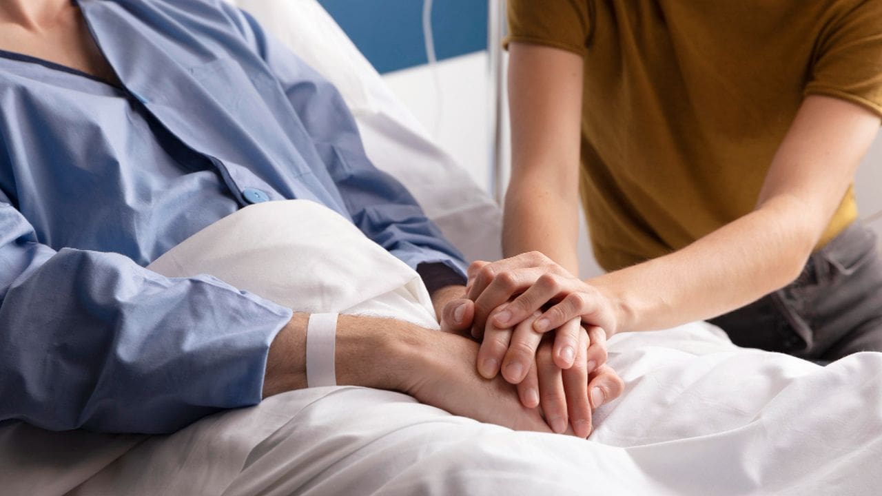 Paciente em leito hospitalar com alguém segurando suas mãos em sinal de apoio.