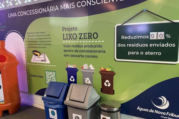 Painel informativo sobre o projeto Lixo Zero na concessionária Águas de Nova Friburgo, com lixeiras de coleta seletiva e um recipiente para descarte de óleo.