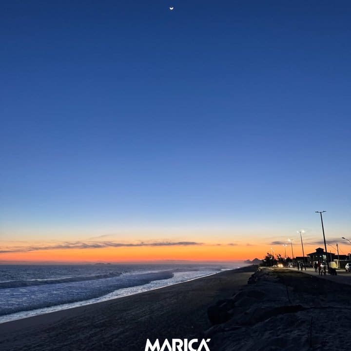 Paisagem da praia de Maricá ao entardecer, com céu azul e lua crescente