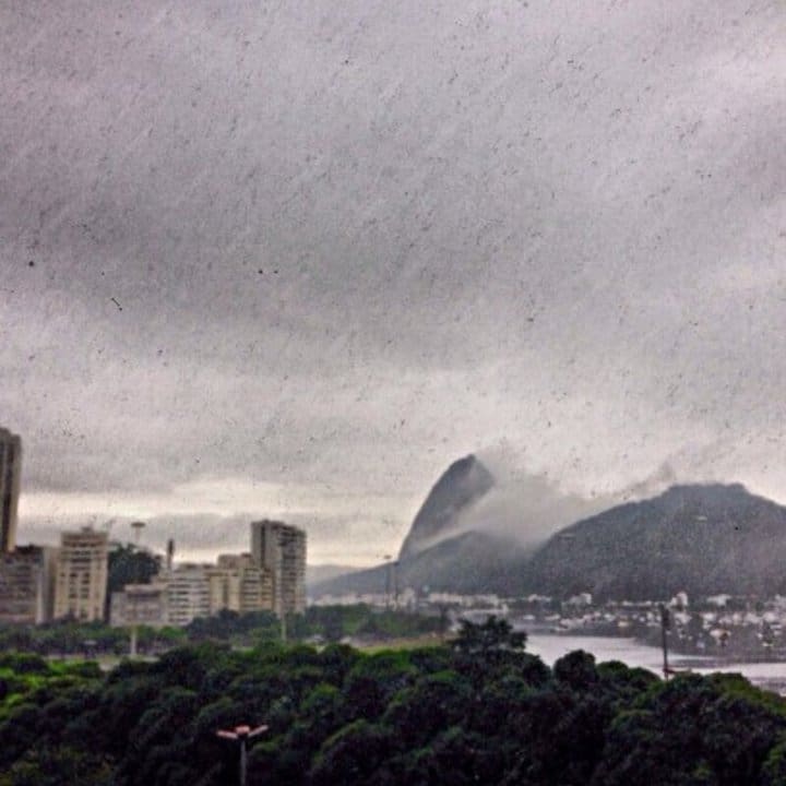 Paisagem urbana do Rio de Janeiro com montanhas e céu nublado. Predominância de prédios e vegetação.