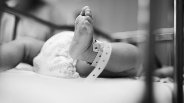 Pé de bebê com pulseira de identificação em ambiente hospitalar, em preto e branco.