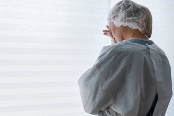 Pessoa em roupa de proteção, possivelmente em ambiente hospitalar, olhando para janela.