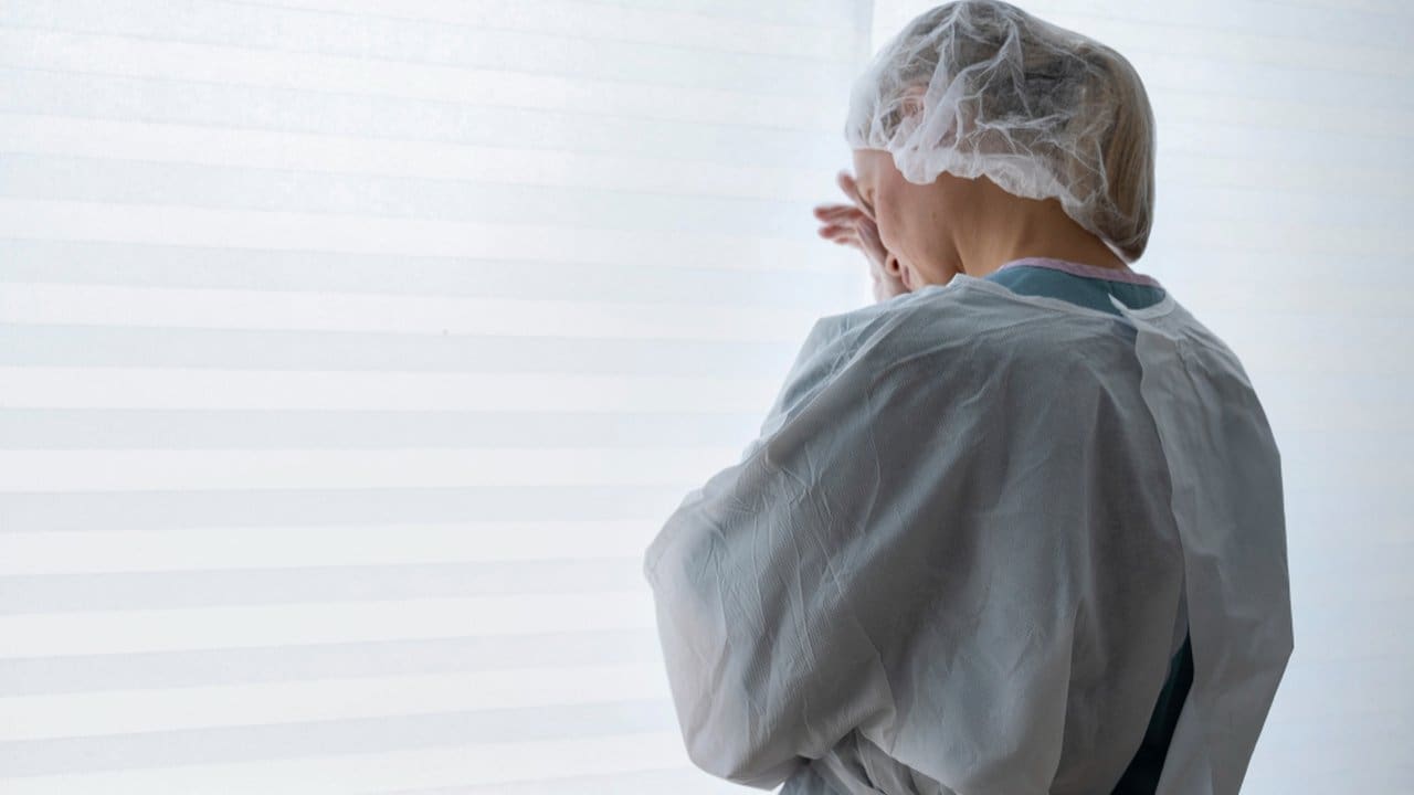 Pessoa em roupa de proteção, possivelmente em ambiente hospitalar, olhando para janela.