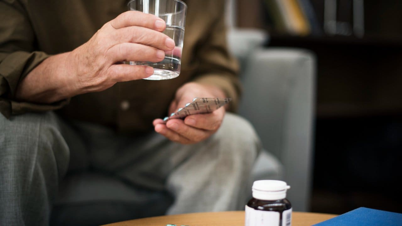 Pessoa segurando um copo d'água e comprimidos, possivelmente tomando medicação para problemas cardíacos.