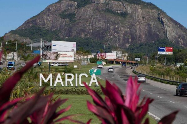 Placa 'Maricá' em frente a rodovia, com montanha ao fundo.