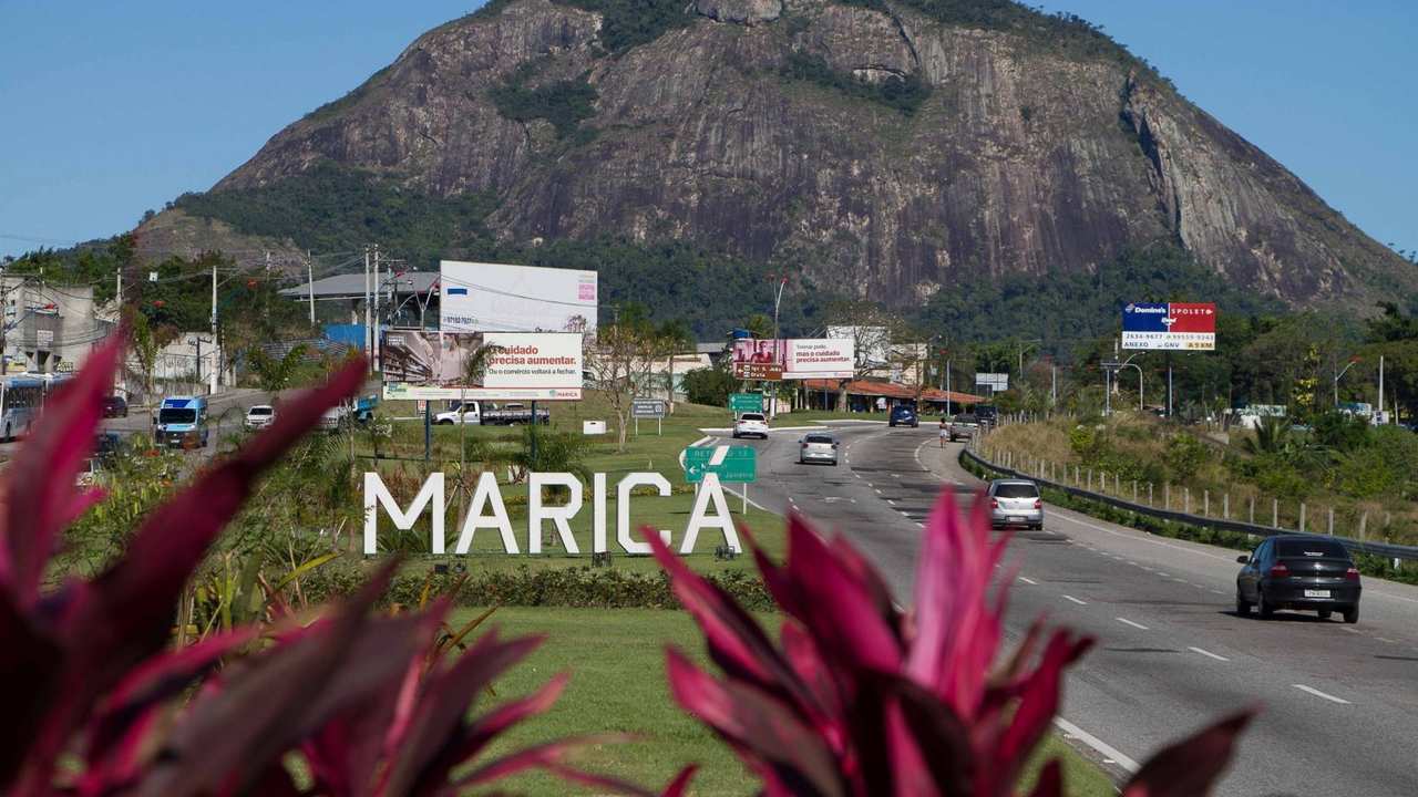 Placa 'Maricá' em frente a rodovia, com montanha ao fundo.