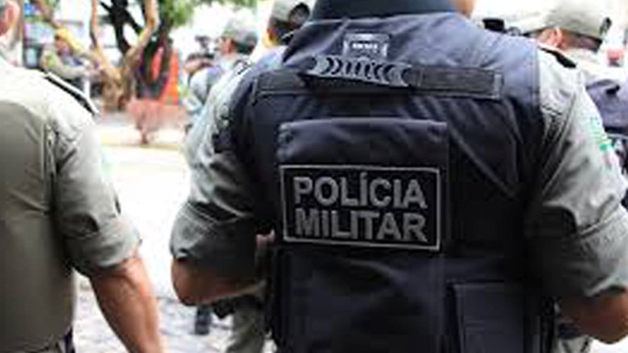 Policial militar com colete tático com a inscrição 'POLÍCIA MILITAR' nas costas.