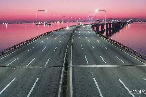 Ponte com tráfego rodoviário e céu rosado ao fundo, com reflexos na água.