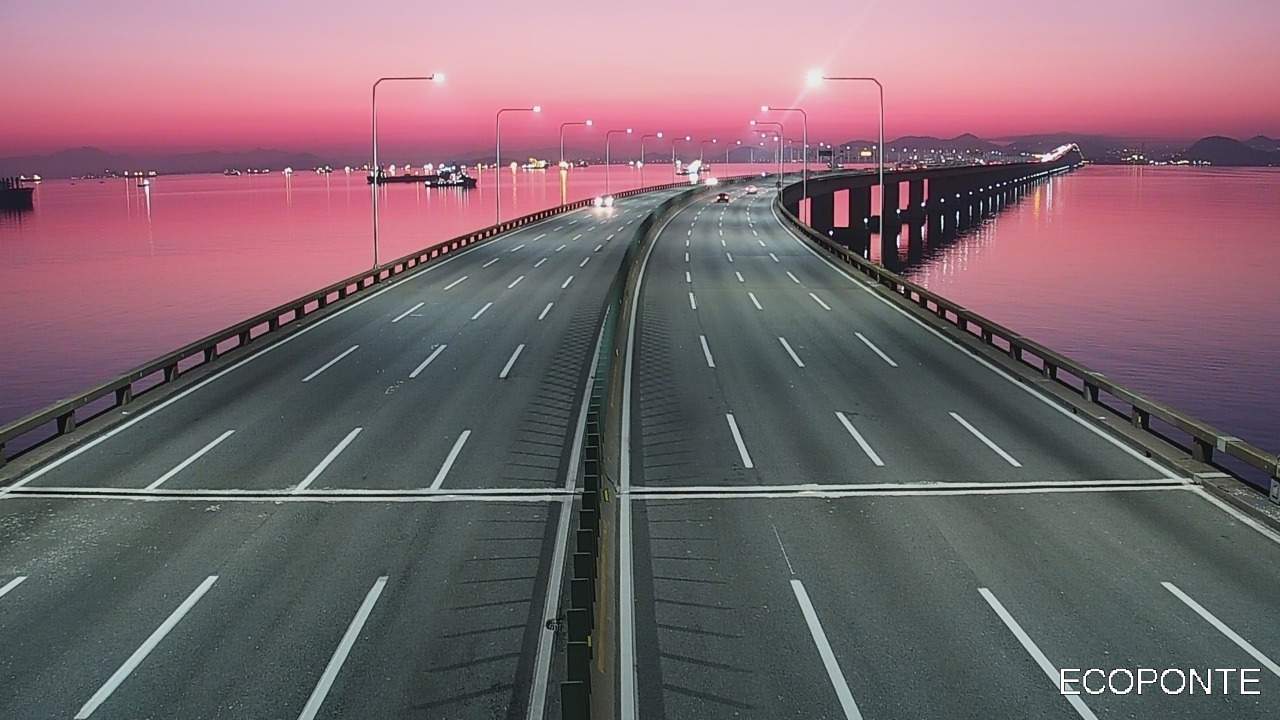 Ponte com tráfego rodoviário e céu rosado ao fundo, com reflexos na água.
