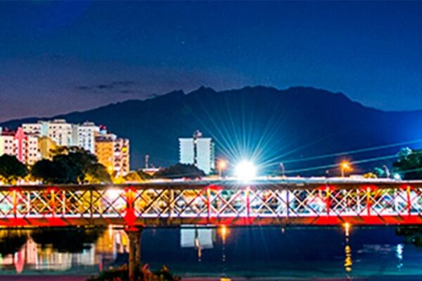 Ponte iluminada sobre o rio, com prédios coloridos ao fundo e montanhas escuras ao anoitecer em Resende.