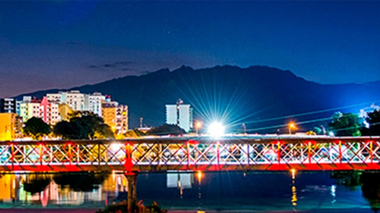 Ponte iluminada sobre o rio, com prédios coloridos ao fundo e montanhas escuras ao anoitecer em Resende.