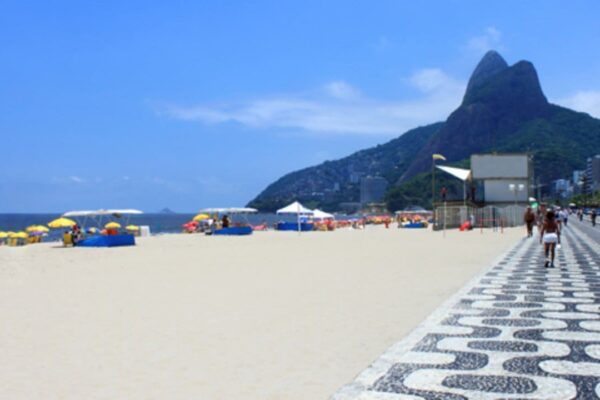 Praia com areia branca e mar azul, com pessoas caminhando na orla e barracas de praia, com uma montanha verde ao fundo.
