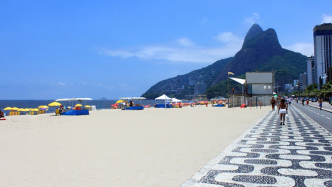 Praia com areia branca e mar azul, com pessoas caminhando na orla e barracas de praia, com uma montanha verde ao fundo.